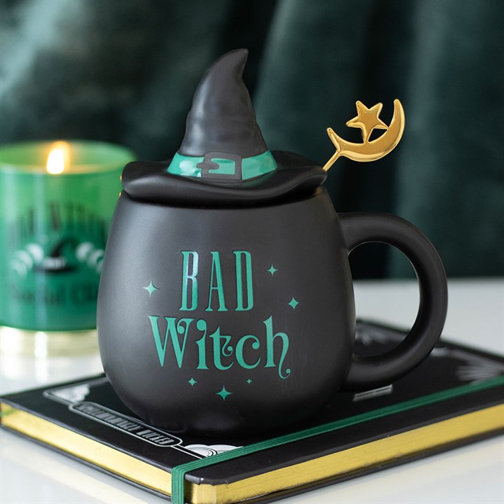 Black mug with witch hat and 'Bad Witch' text, candle, and book in background
