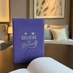 Purple notebook with 'Believe in Yourself' text on a wooden table in a cozy room.