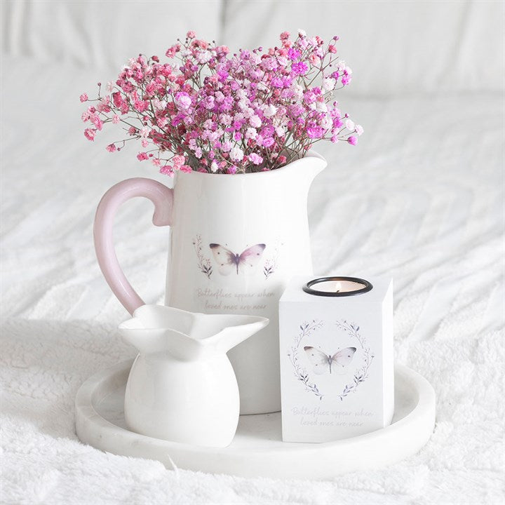 White pitcher with pink flowers and a white candle with butterfly design on a white surface