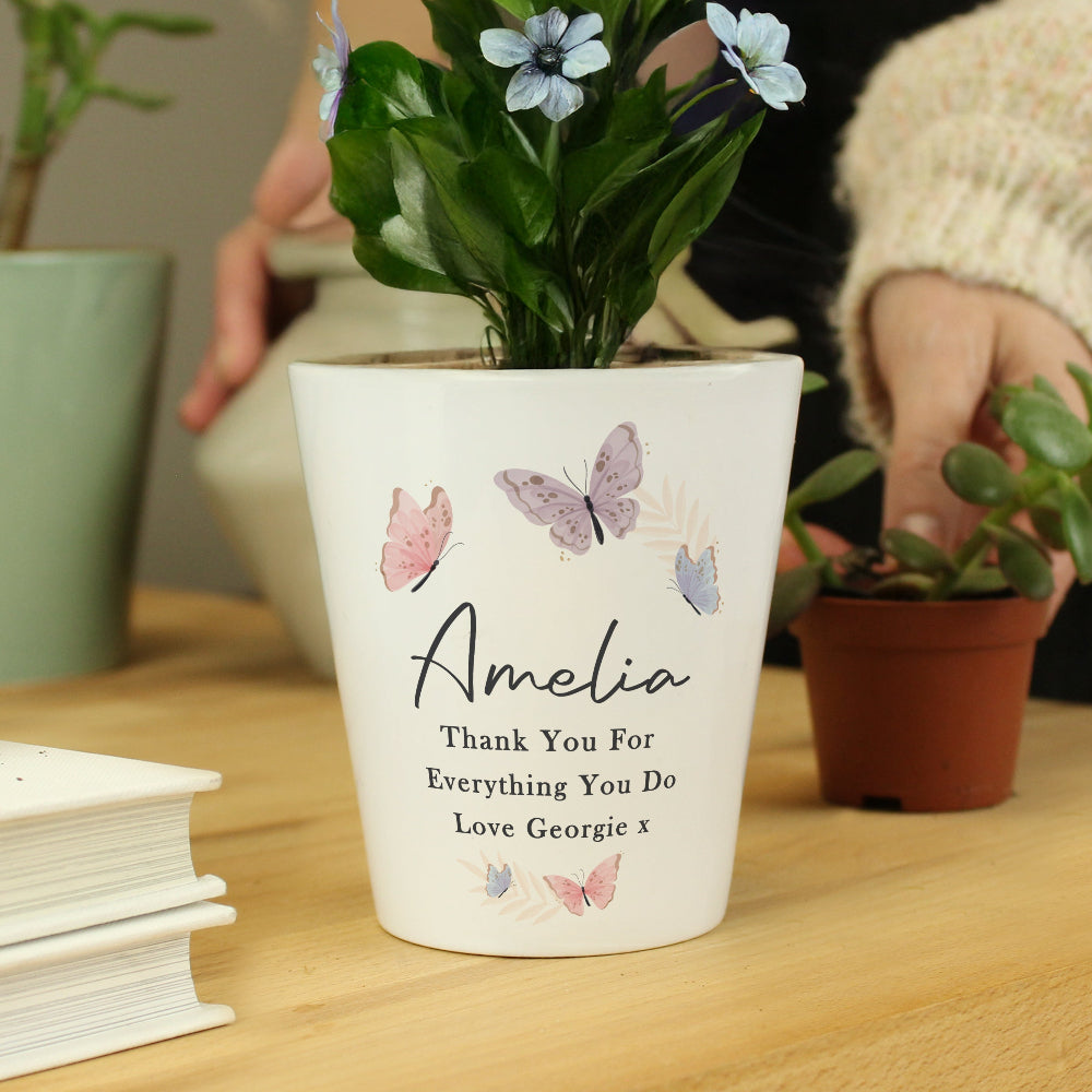 White pot with butterfly design and personalized message on a wooden surface with plants in the background.