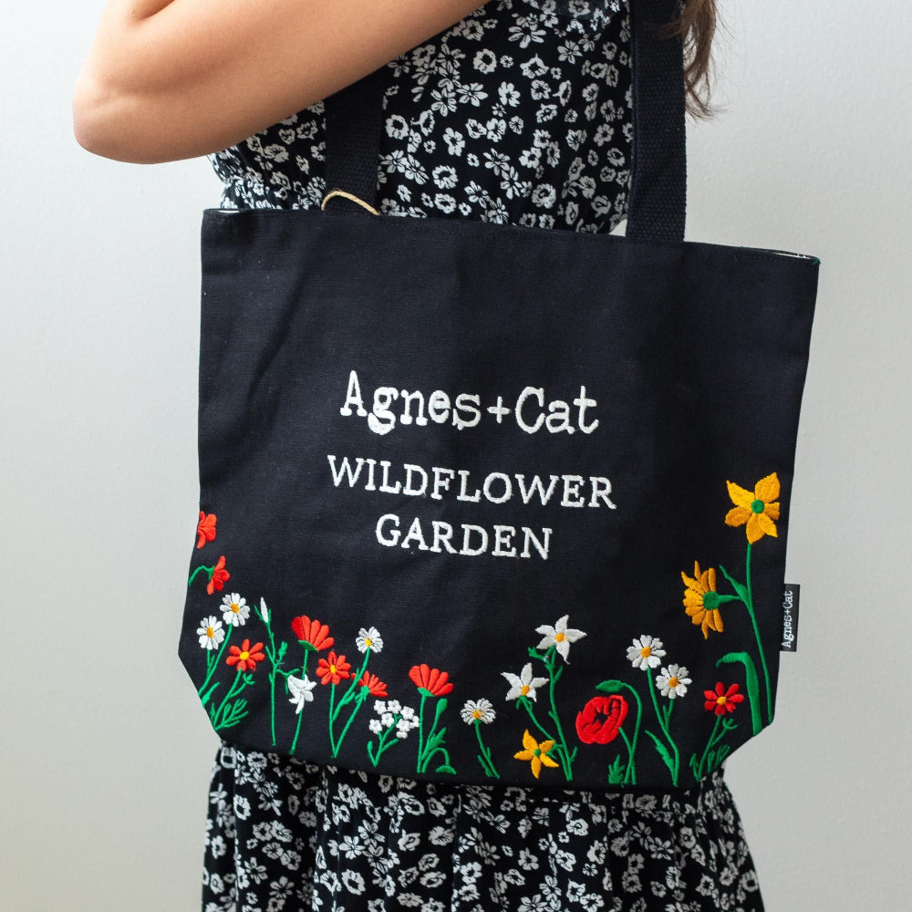Person holding a black tote bag with floral designs and 'Agnes + Cat Wildflower Garden' text.