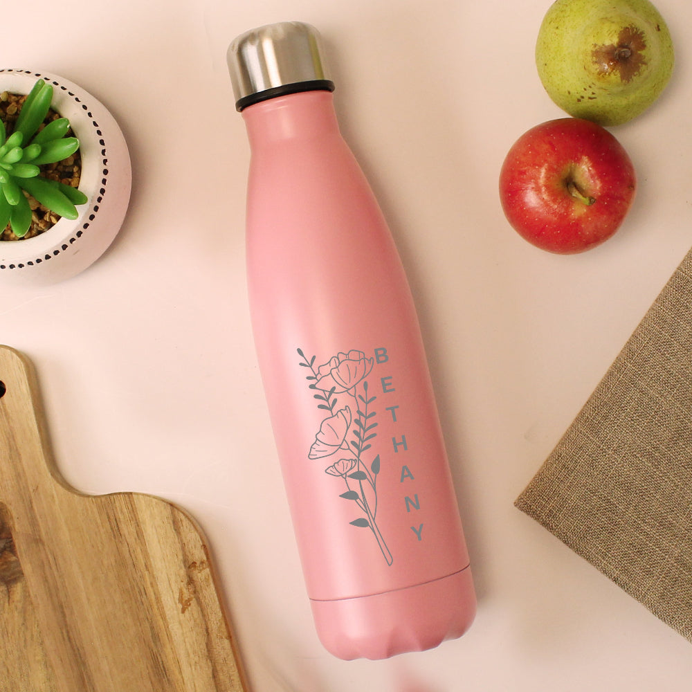 Pink water bottle with floral design and 'BETHANY' text on a light surface with apples and a plant.