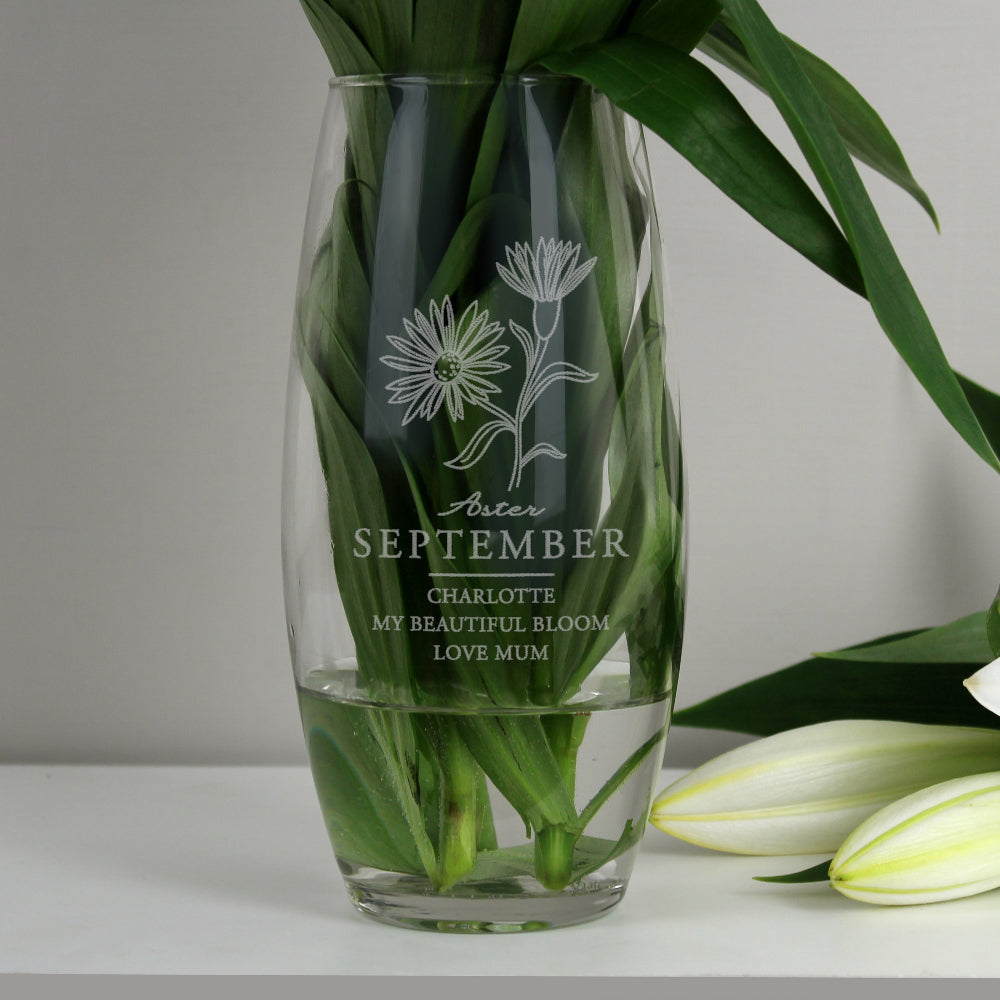 Clear glass vase with engraved floral design and text, filled with water and green leaves on a white surface.