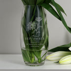 Clear glass vase with engraved floral design and text, filled with water and green leaves on a white surface.
