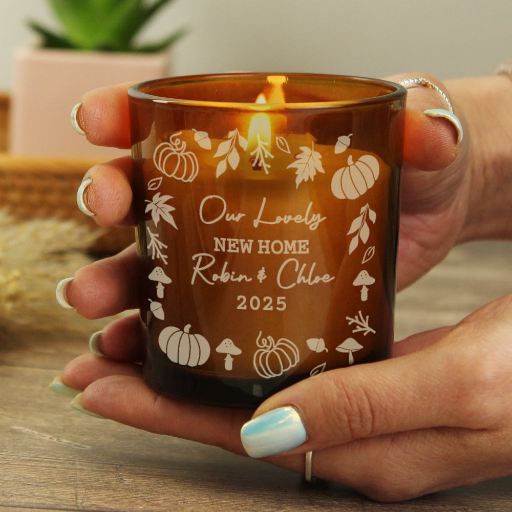 Person holding a lit candle in an amber glass holder with engraved text and autumn designs.