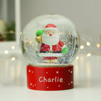 Snow globe with Santa Claus figure on a red base with 'Charlie' written on it, against a blurred light background.