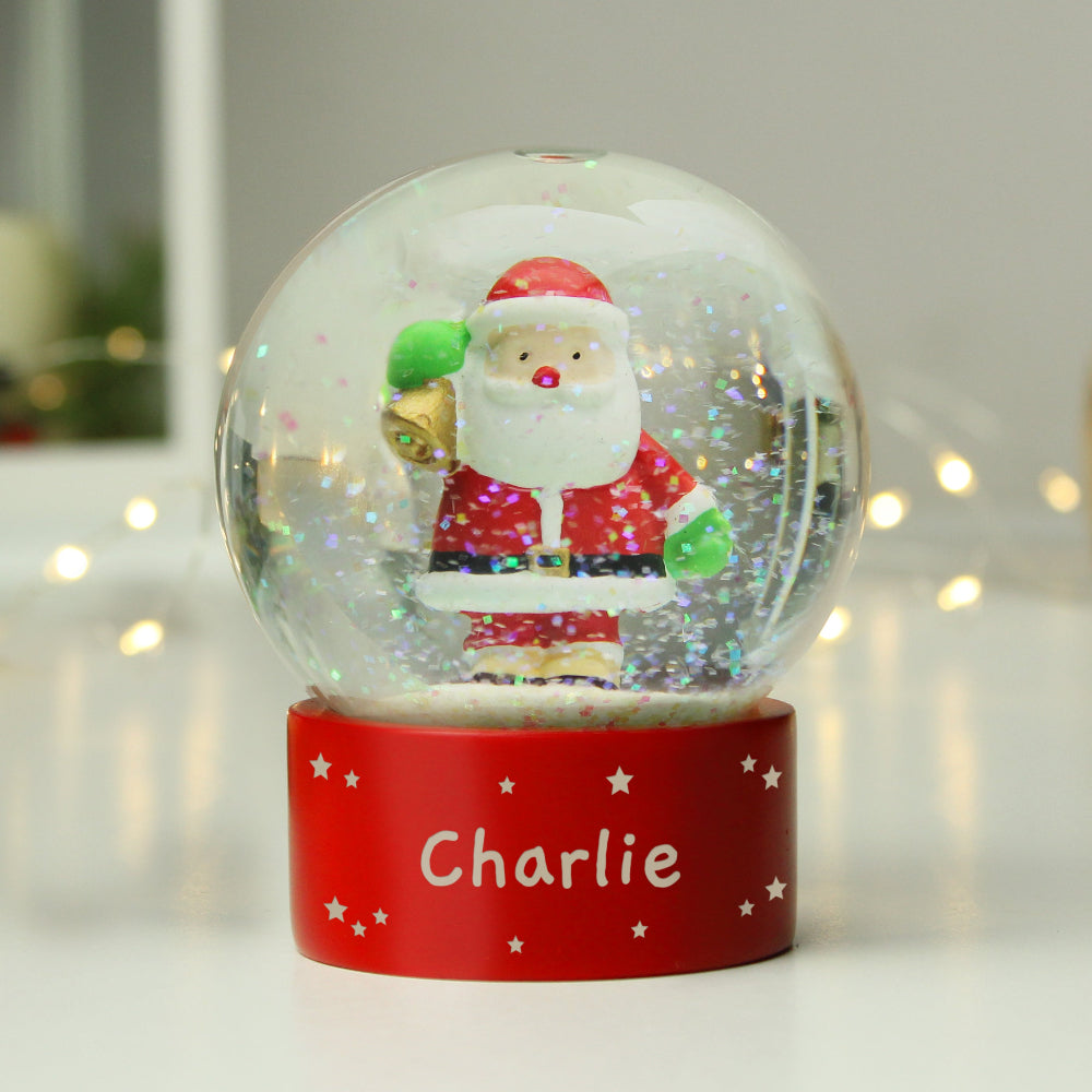 Snow globe with Santa Claus figure on a red base with 'Charlie' written on it, against a blurred light background.