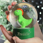 Green dinosaur toy inside a snow globe with 'Freddie' on a green base, held by a person.