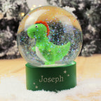 Snow globe with a green dinosaur figure inside, on a wooden surface with a blurred Christmas tree background.