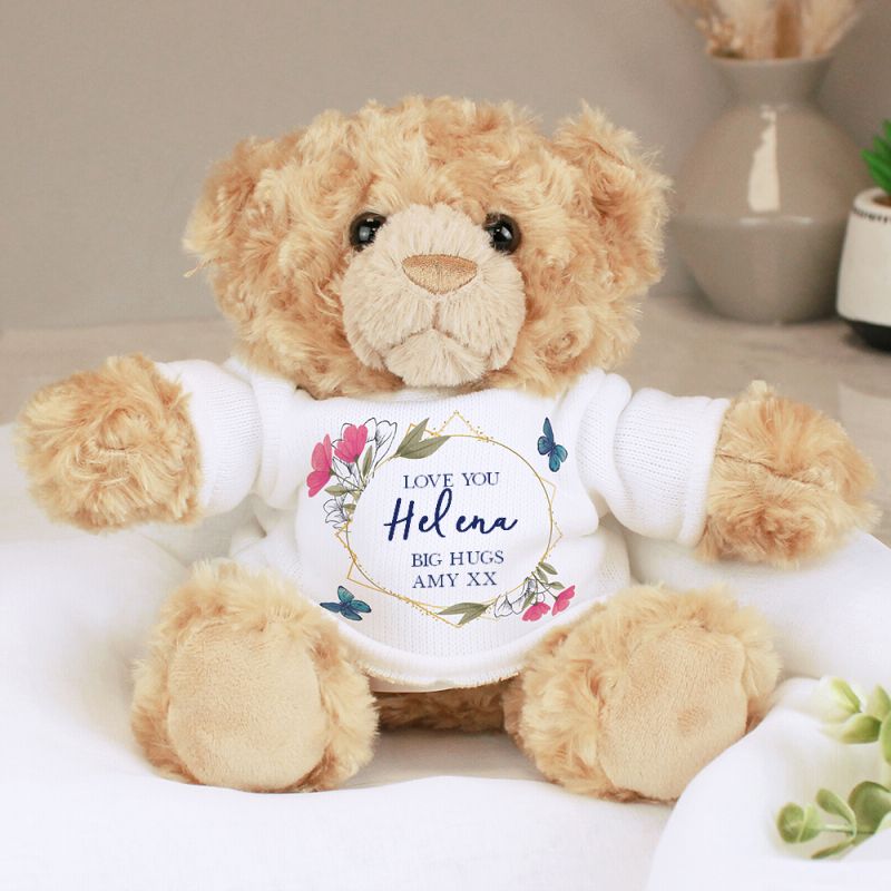 Teddy bear wearing a white shirt with floral design and text on a neutral background