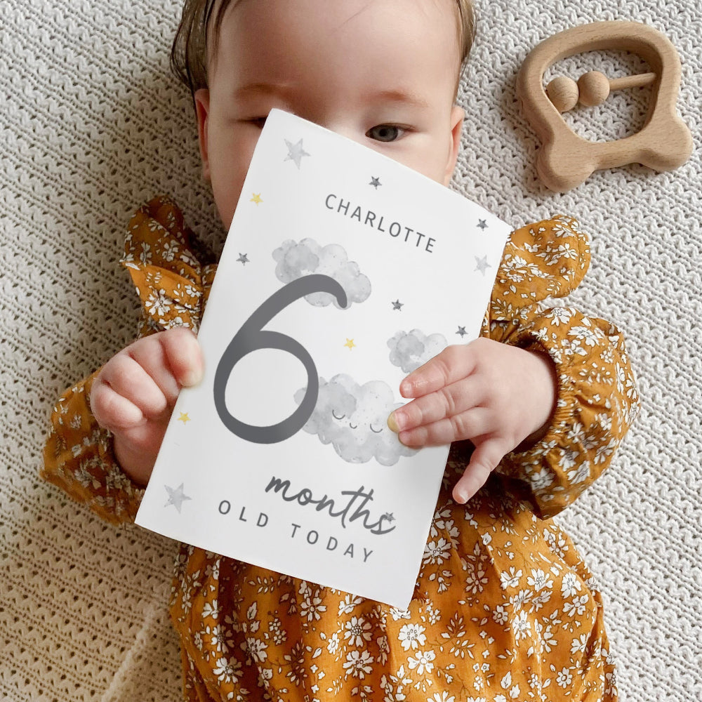 Baby holding a card saying '6 months old today' with a wooden teething ring in the background.