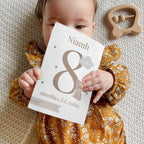 Baby holding a milestone card with 'Niamh 8 months old today' on a textured surface.