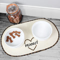 Pet feeding area with two bowls, a heart-shaped mat, and a jar of treats on a wooden floor.