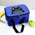 Blue lunch bag with gaming controller design and 'THOMAS' text, placed on a white surface with a banana and green apple.