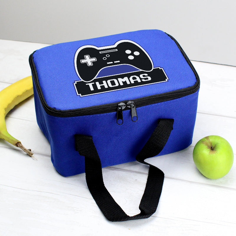 Blue lunch bag with gaming controller design and 'THOMAS' text, placed on a white surface with a banana and green apple.