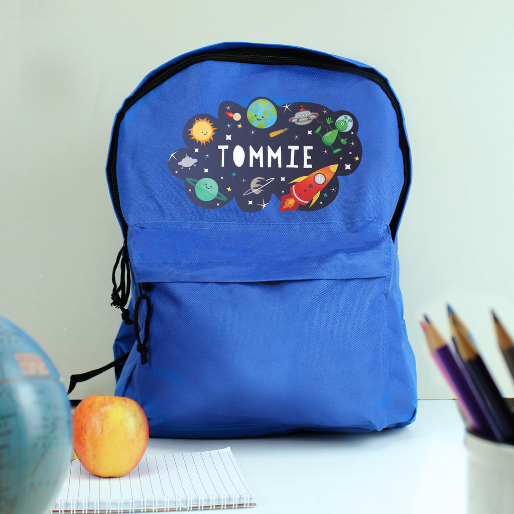 Blue backpack with space-themed design and 'Tommie' text, placed on a surface with stationery items.