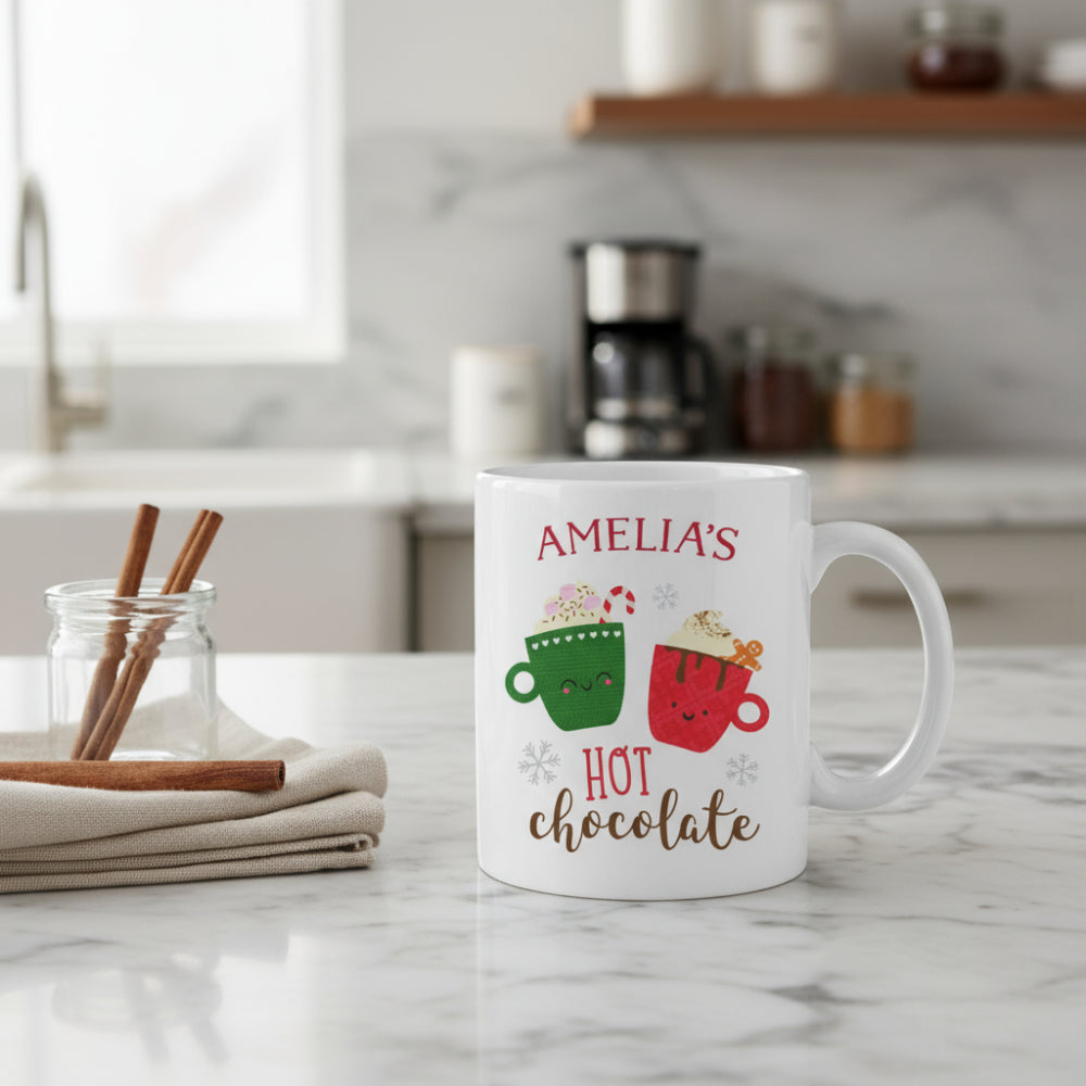White mug with 'AMELIA'S HOT chocolate' text and cartoon mugs on a wooden surface with Christmas decorations.