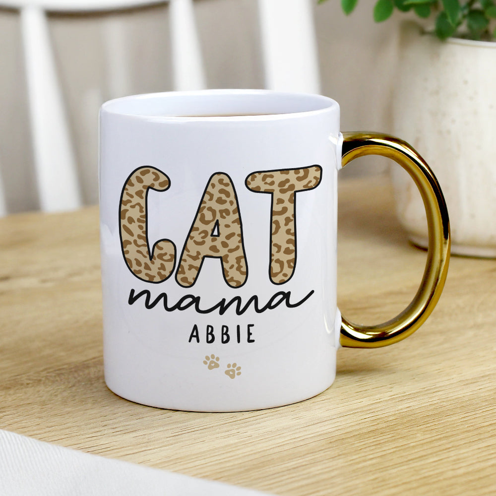 White mug with gold handle featuring 'CAT mama ABBIE' text on a wooden surface.