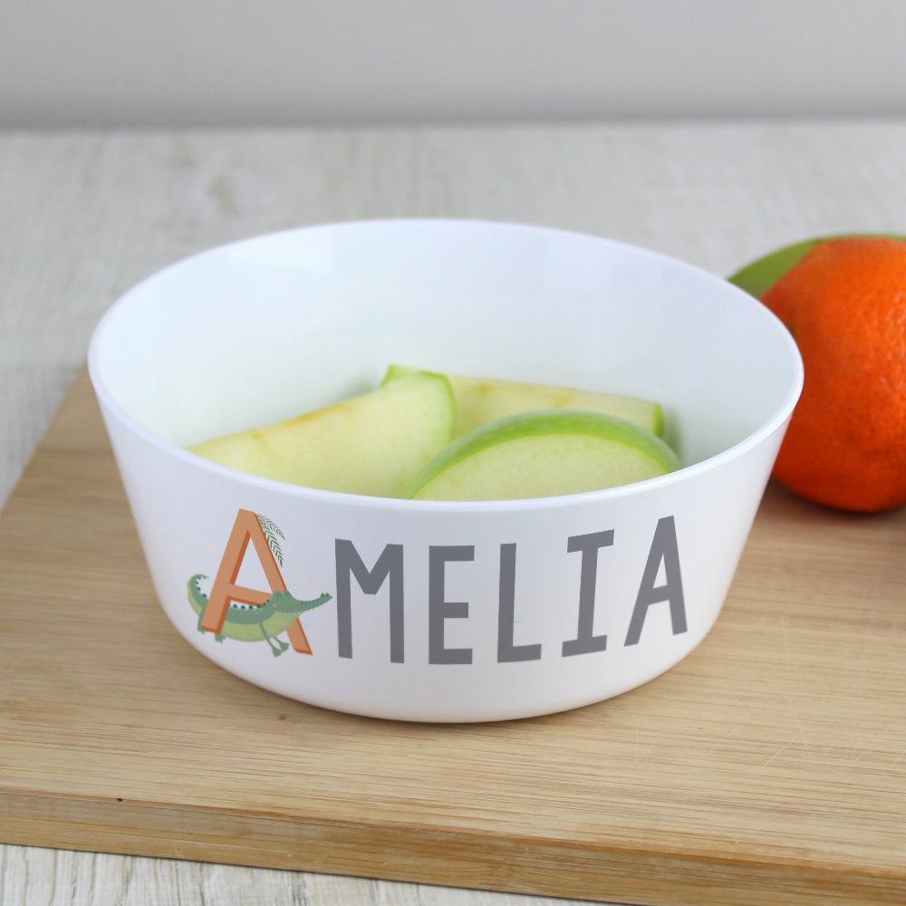 White bowl with 'AMELIA' and a crocodile design on a wooden surface with fruit.
