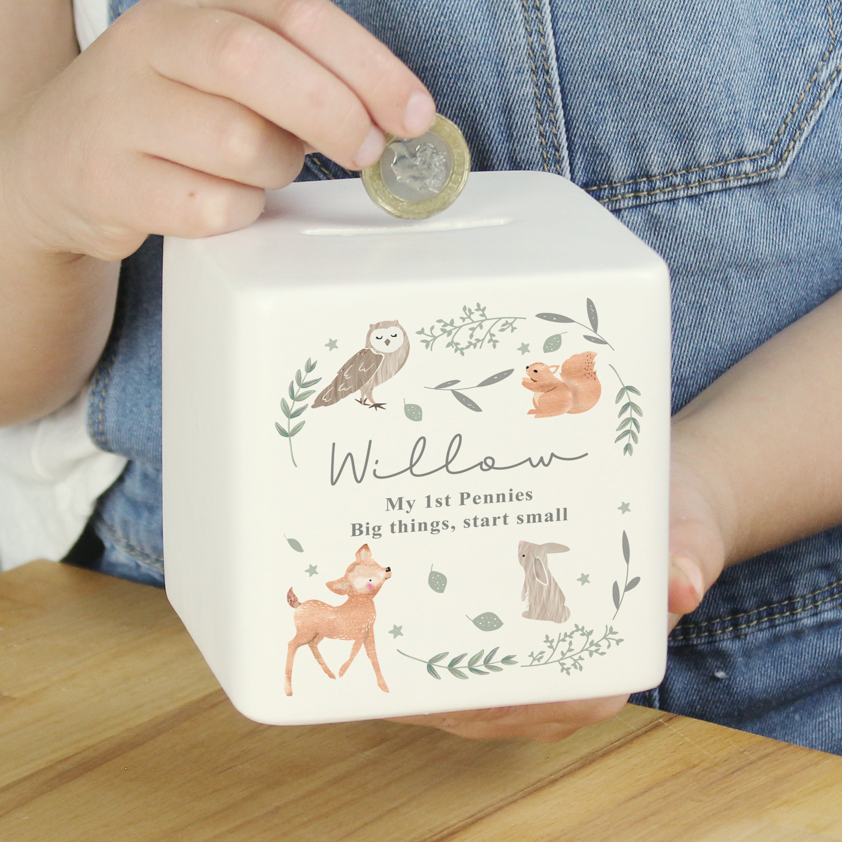 Child's hand placing a coin into a white piggy bank with animal illustrations and text.