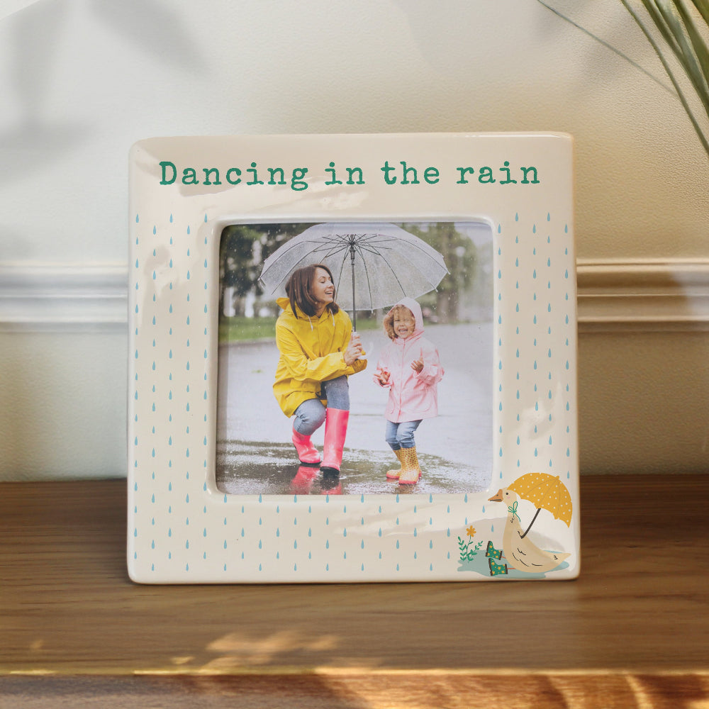 Decorative photo frame with a picture of two children in raincoats and boots, text 'Dancing in the rain', and decorative elements on a wooden surface.