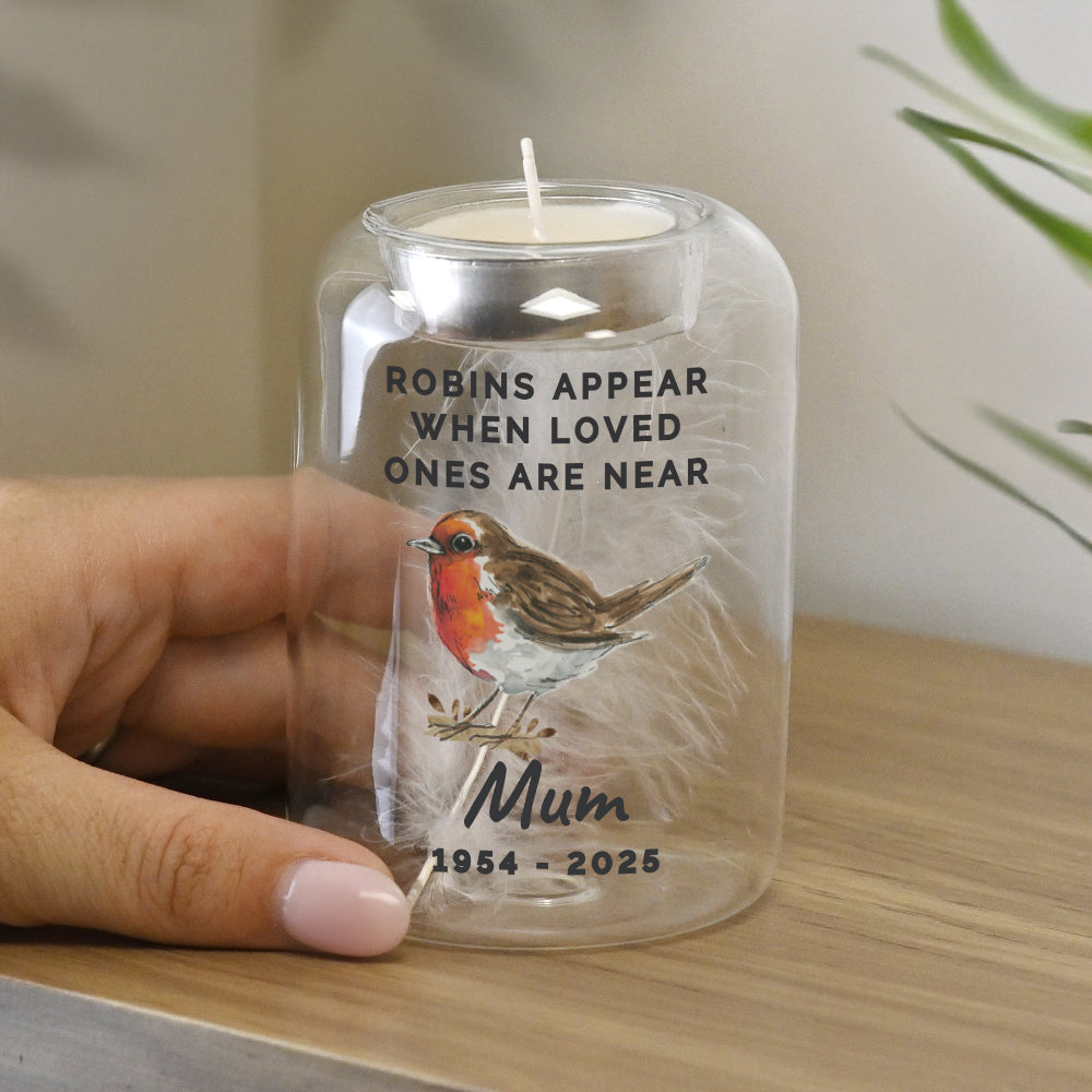 Candle in a glass jar with a robin design and text, held by a hand on a wooden surface.