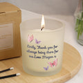 Candle with butterflies and text on a wooden coaster, next to a cardboard box and matches.