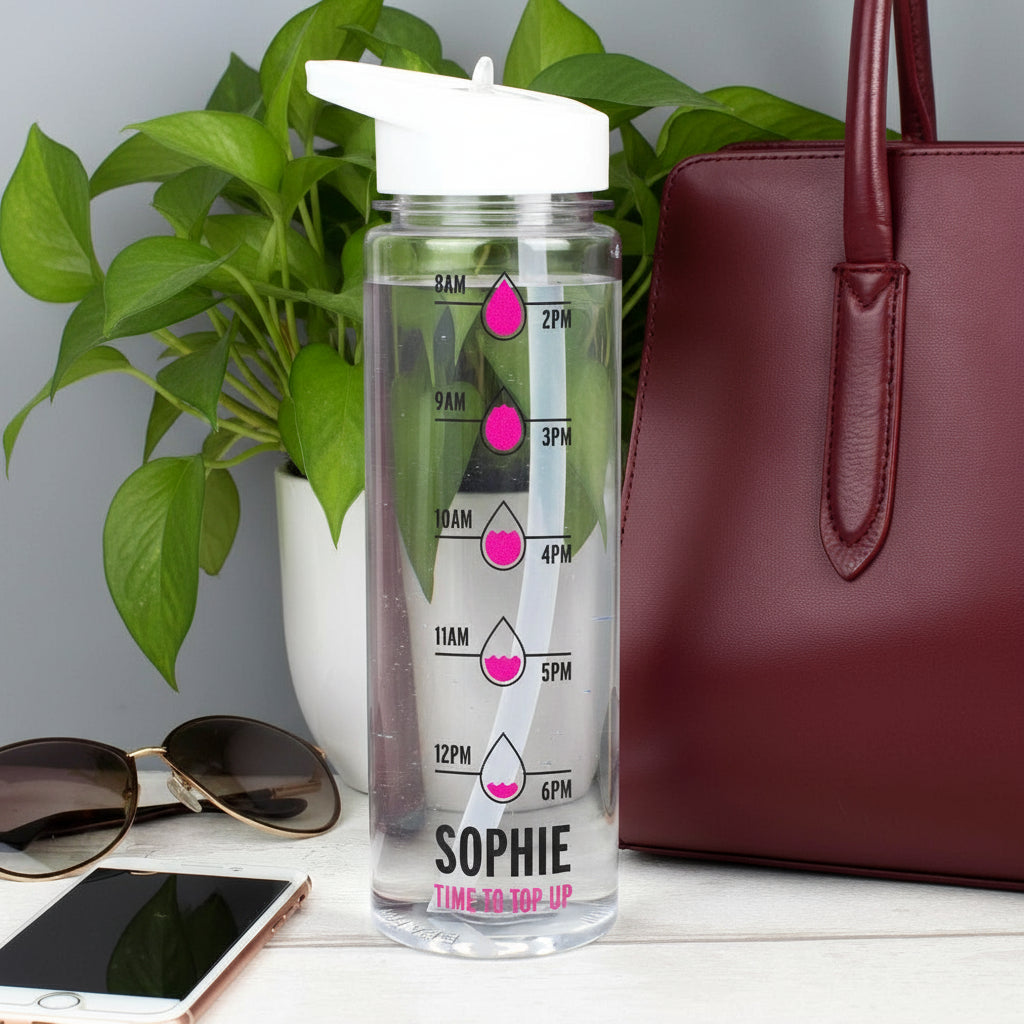 Clear water bottle with time markers and 'SOPHIE' branding, next to a maroon handbag and sunglasses on a light surface.