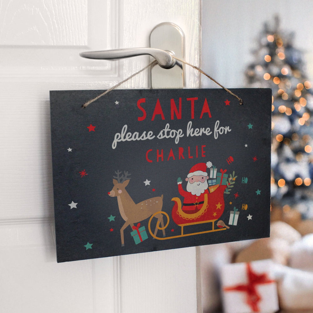 Christmas-themed sign with Santa and reindeer, hanging on a door with a blurred Christmas tree in the background.