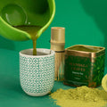 Green tea being poured into a cup with matcha powder and packaging on a green background