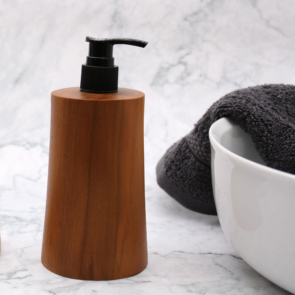 Wooden soap dispenser with black pump on a marble surface with a towel and bowl in the background.