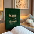 Green notebook with 'Make It Happen' text on a table in a cozy room.