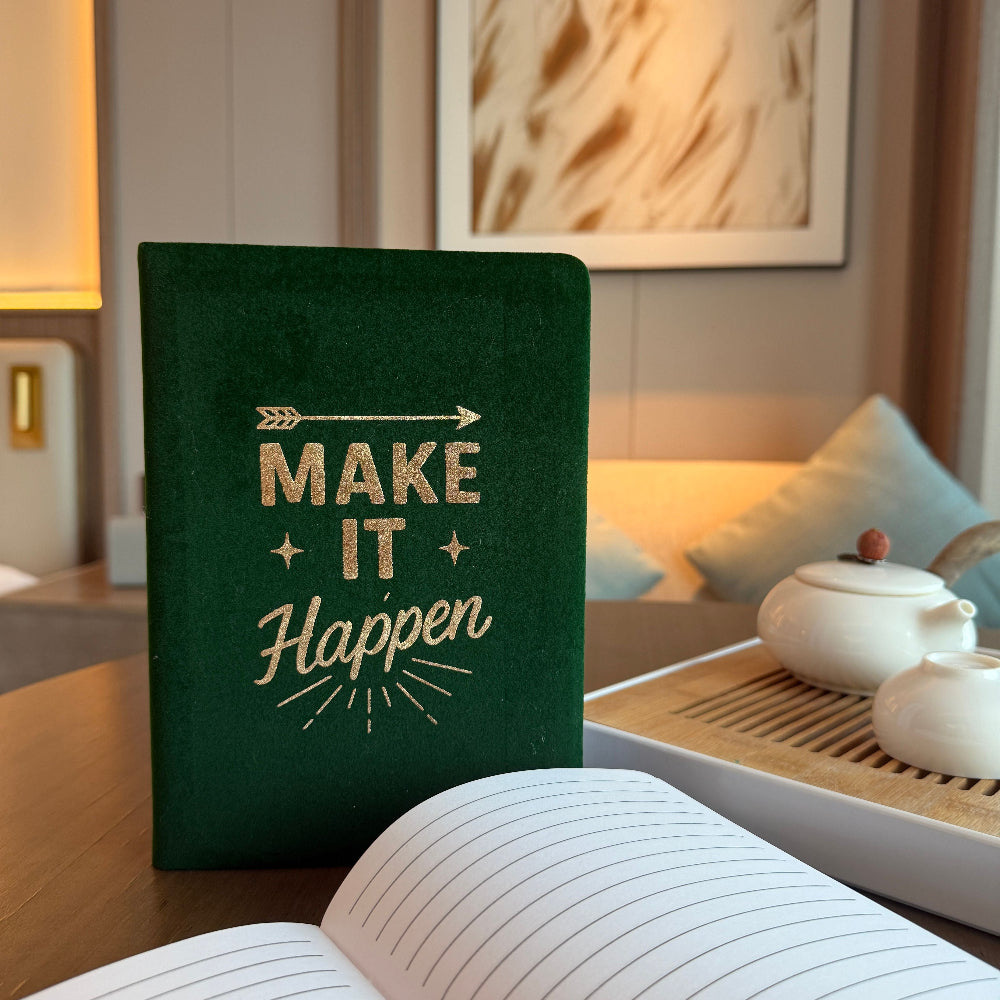Green notebook with 'Make It Happen' text on a table in a cozy room.