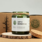 Candle labeled 'Ash & Woodsmoke' with a green label and wooden lid, placed on a wooden surface with a cardboard box in the background.