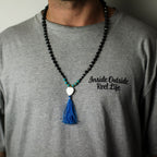 Person wearing a gray t-shirt with text and a beaded necklace with a blue tassel.
