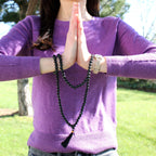 Person wearing a purple sweater with hands in prayer position, holding black beaded bracelets outdoors.