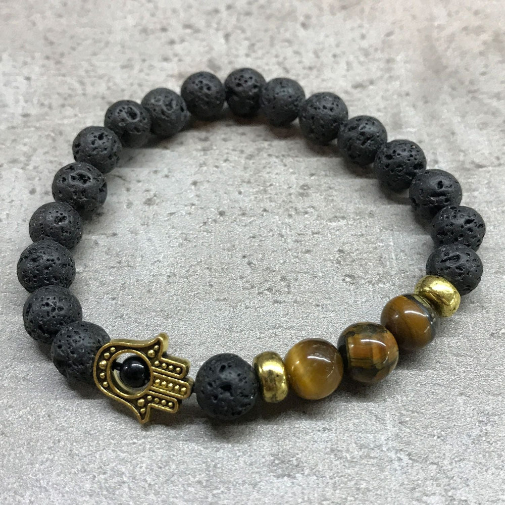 Bracelet with black lava beads, gold Hamsa hand charm, and brown tiger eye stones on a light gray background