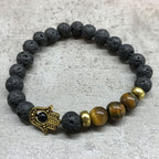 Bracelet with black lava beads, gold Hamsa hand charm, and brown tiger eye stones on a light gray background
