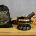 Tibetan singing bowl with wooden striker on a wooden surface, with a decorative bag in the background.