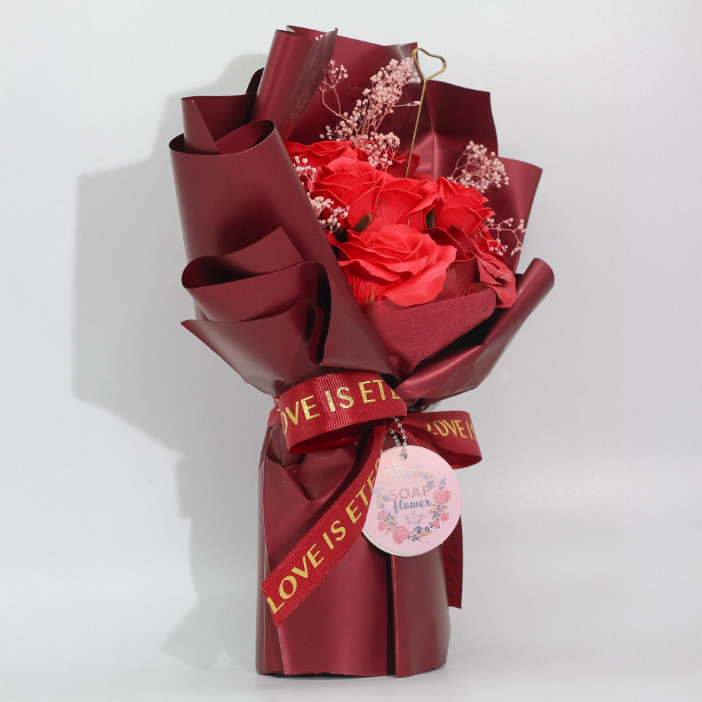Bouquet of soap roses wrapped in red paper with 'LOVE IS' ribbon on a white background