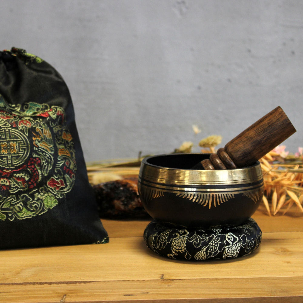 Decorative black and gold singing bowl with wooden striker on a wooden surface, with a patterned bag in the background.