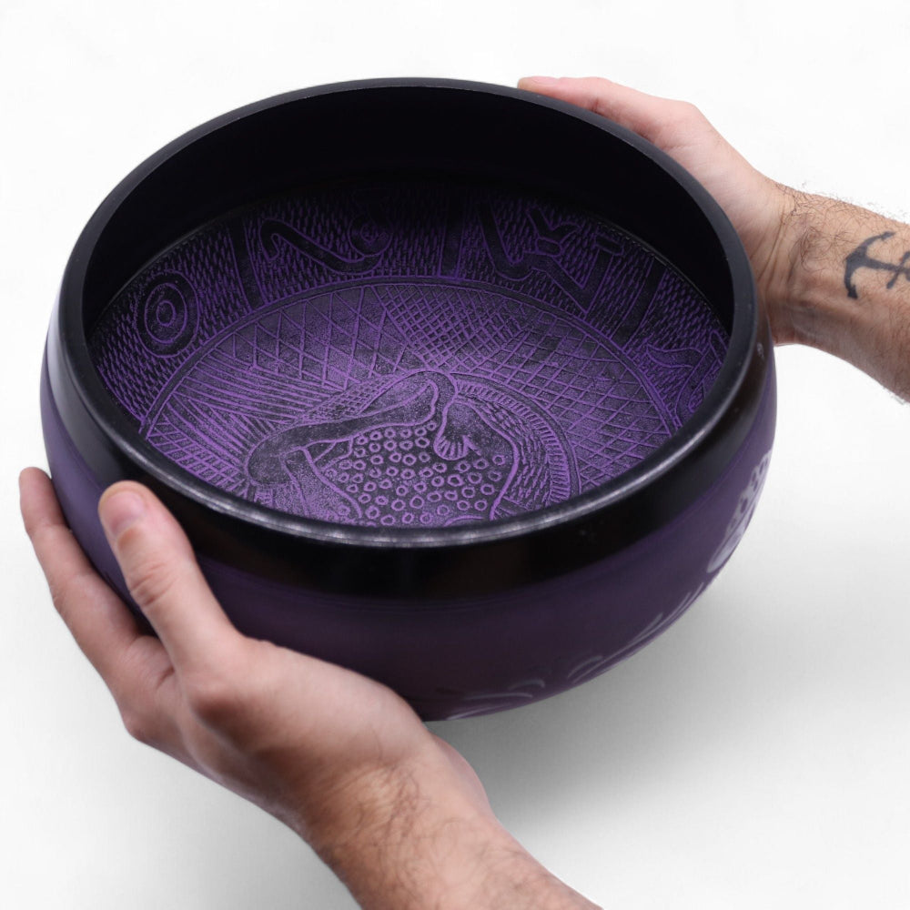 Purple singing bowl with intricate designs held by hands on a white background