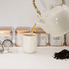 Tea being poured from a white teapot into a white mug with tea packaging in the background.