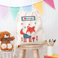 Children's drawstring bag with fox design and name 'Martha', teddy bear, and stationery on a wooden stool.