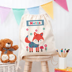 Children's drawstring bag with fox design and name 'Martha', teddy bear, and stationery on a wooden stool.