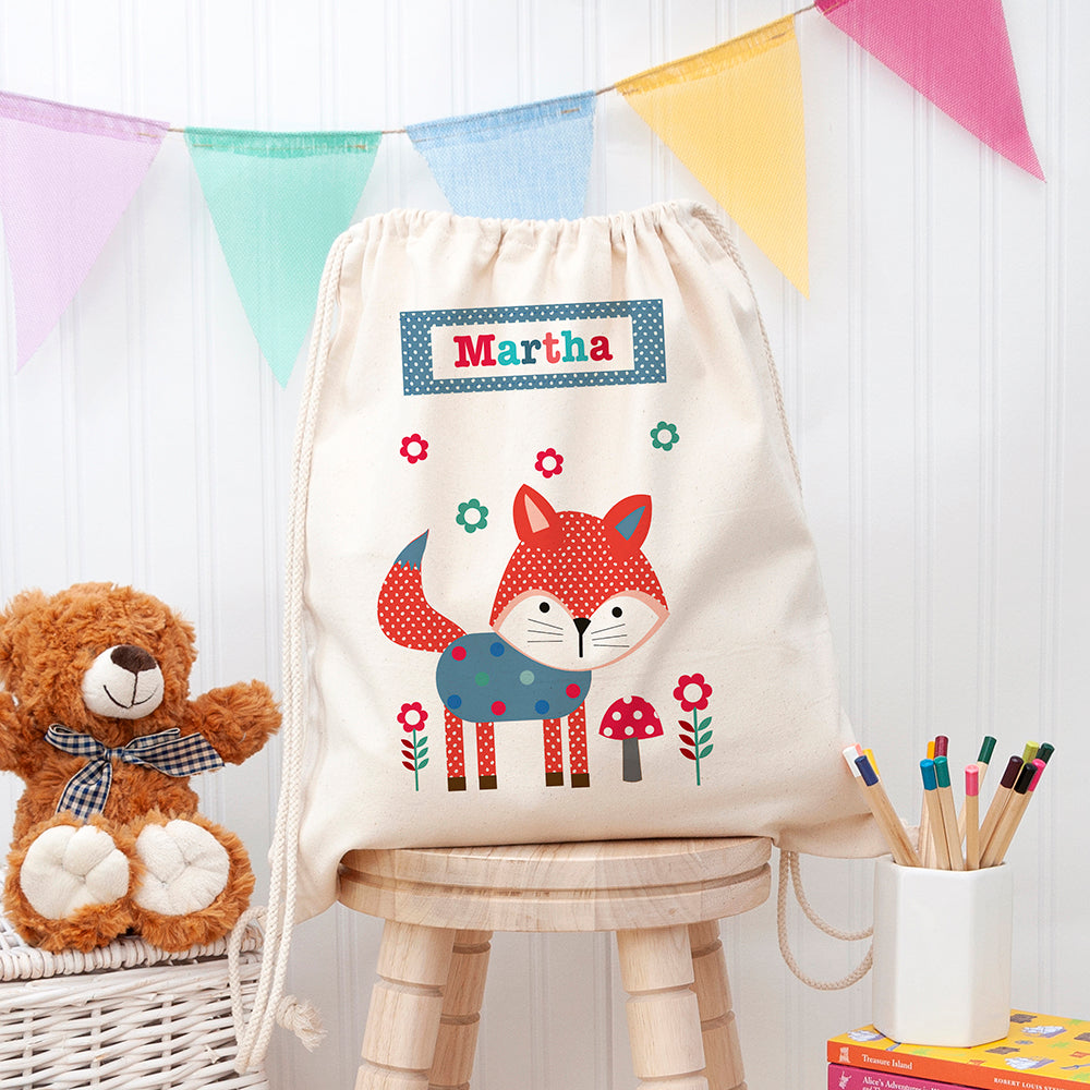 Children's drawstring bag with fox design and name 'Martha', teddy bear, and stationery on a wooden stool.